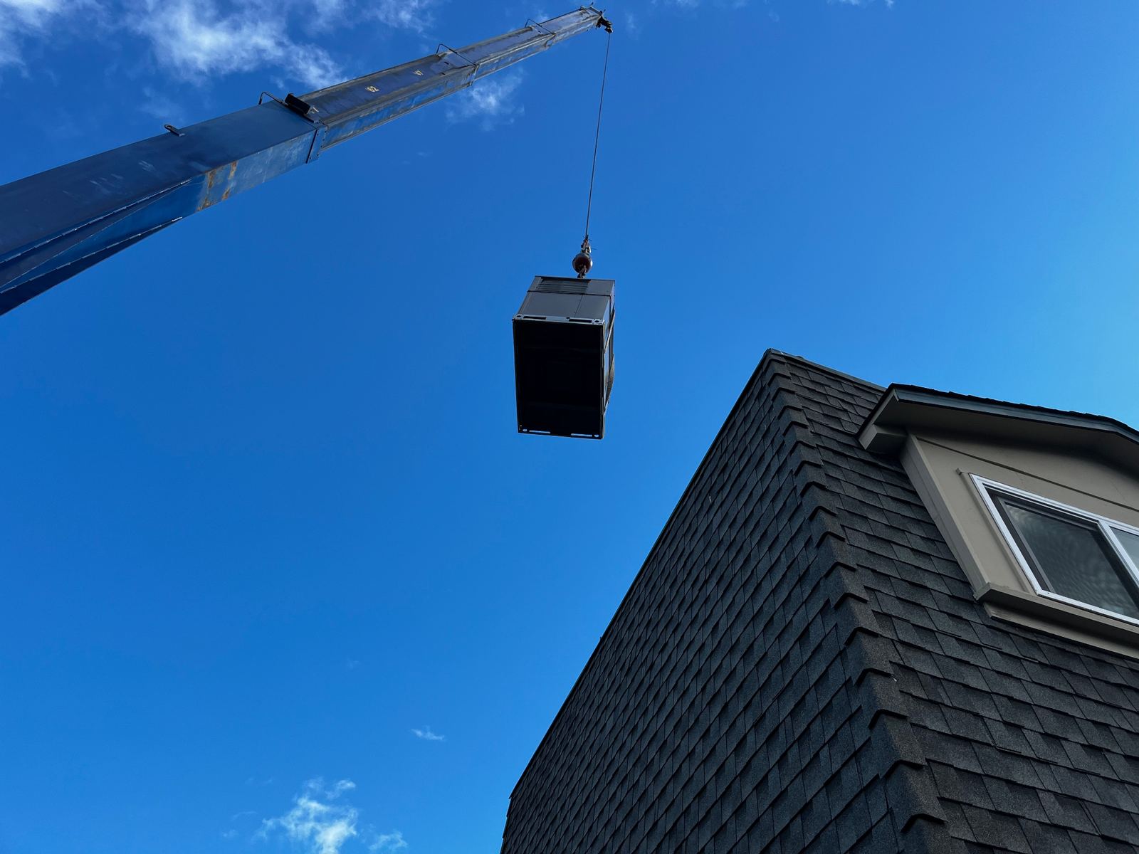 Crane lifting a new rooftop packaged HVAC unit onto a residential roof against a clear blue sky.