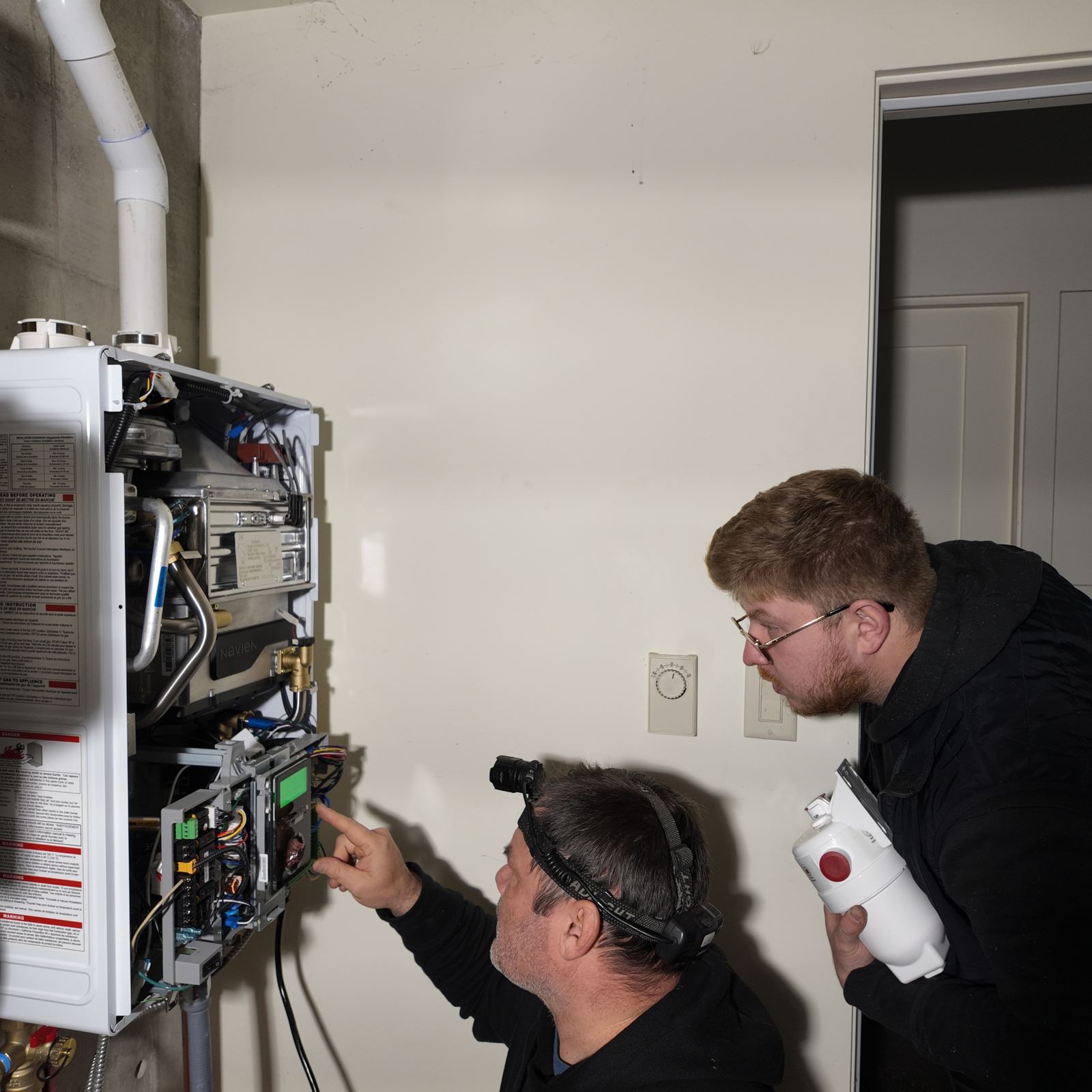 ABC Service technicians diagnosing a Navien condensing tankless water heater control board with a headlamp during a Bay Area service call.